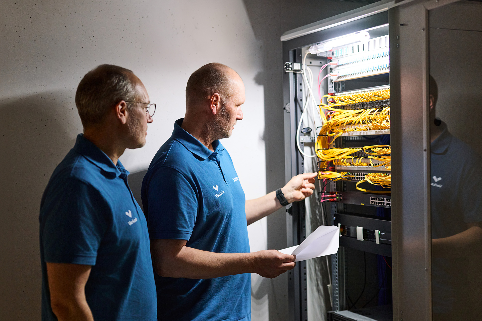 Zwei Männer in blauen Poloshirts prüfen einen Serverrack mit gelben Netzwerkkabeln in einem Technikraum.