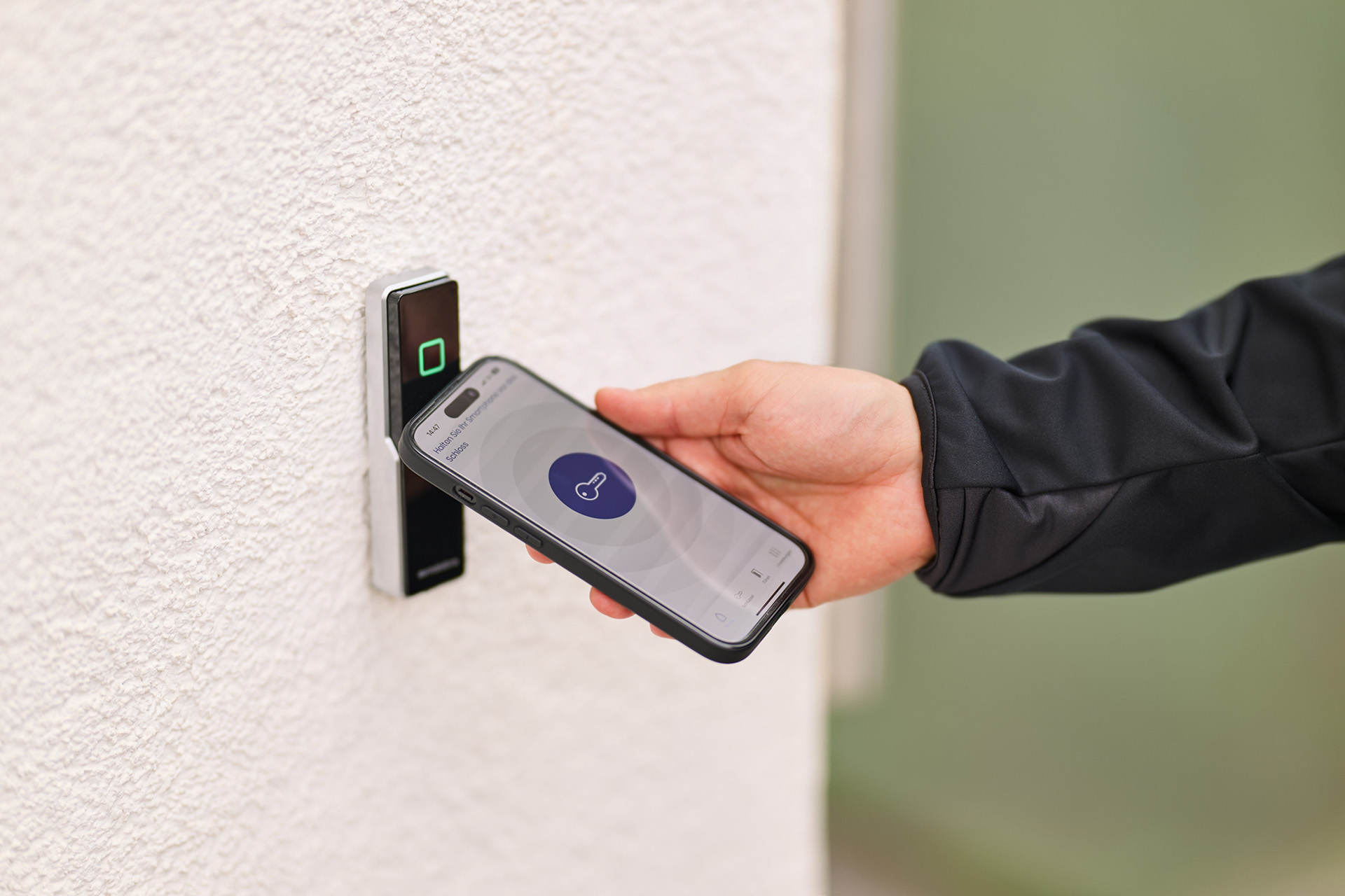 Hand hält Smartphone mit geöffneter App vor elektronisches Türschloss mit grün leuchtendem Quadrat.