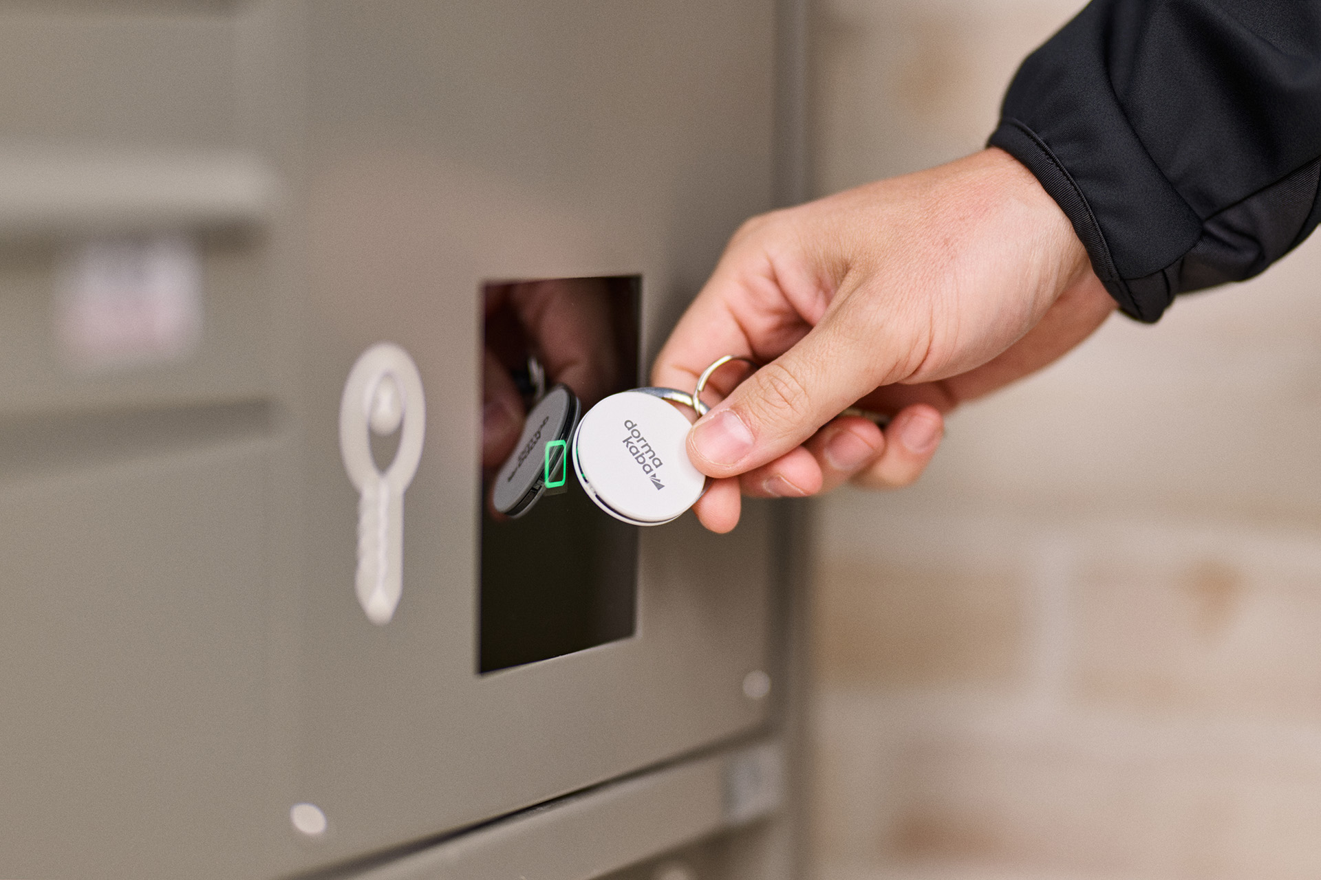 Hand hält weißen Schlüsselanhänger mit Aufschrift 'dormakaba' vor schwarzen Kartenleser an grauem Schließfach