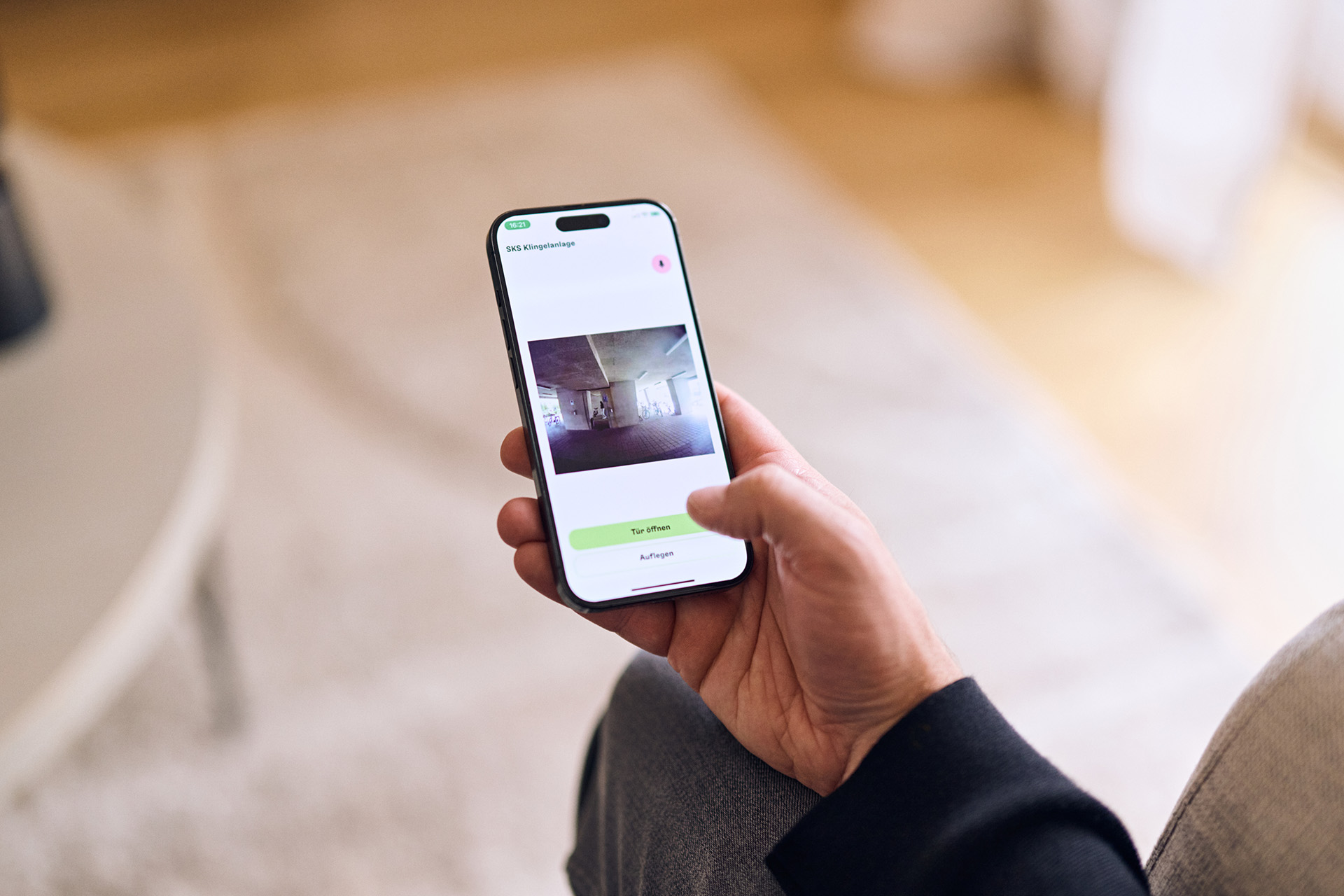 Hand hält Smartphone mit Anzeige einer Raumüberwachung und den Schaltflächen 'Zur Aufnahme' und 'Auflegen' auf dem Bildschirm.