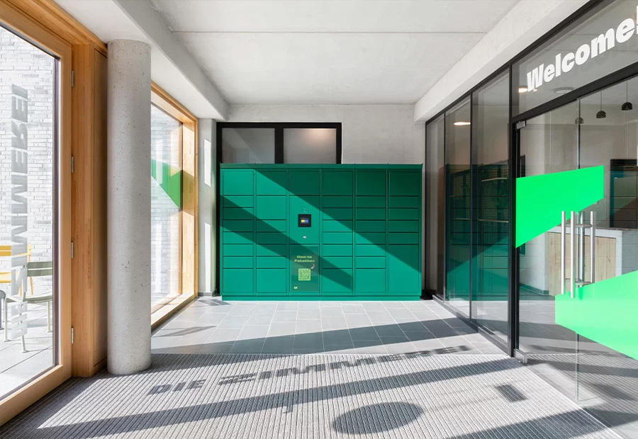 Grüne Paketstation mit mehreren Fächern in einem hellen Eingangsbereich mit Glaswänden und Holzrahmen.