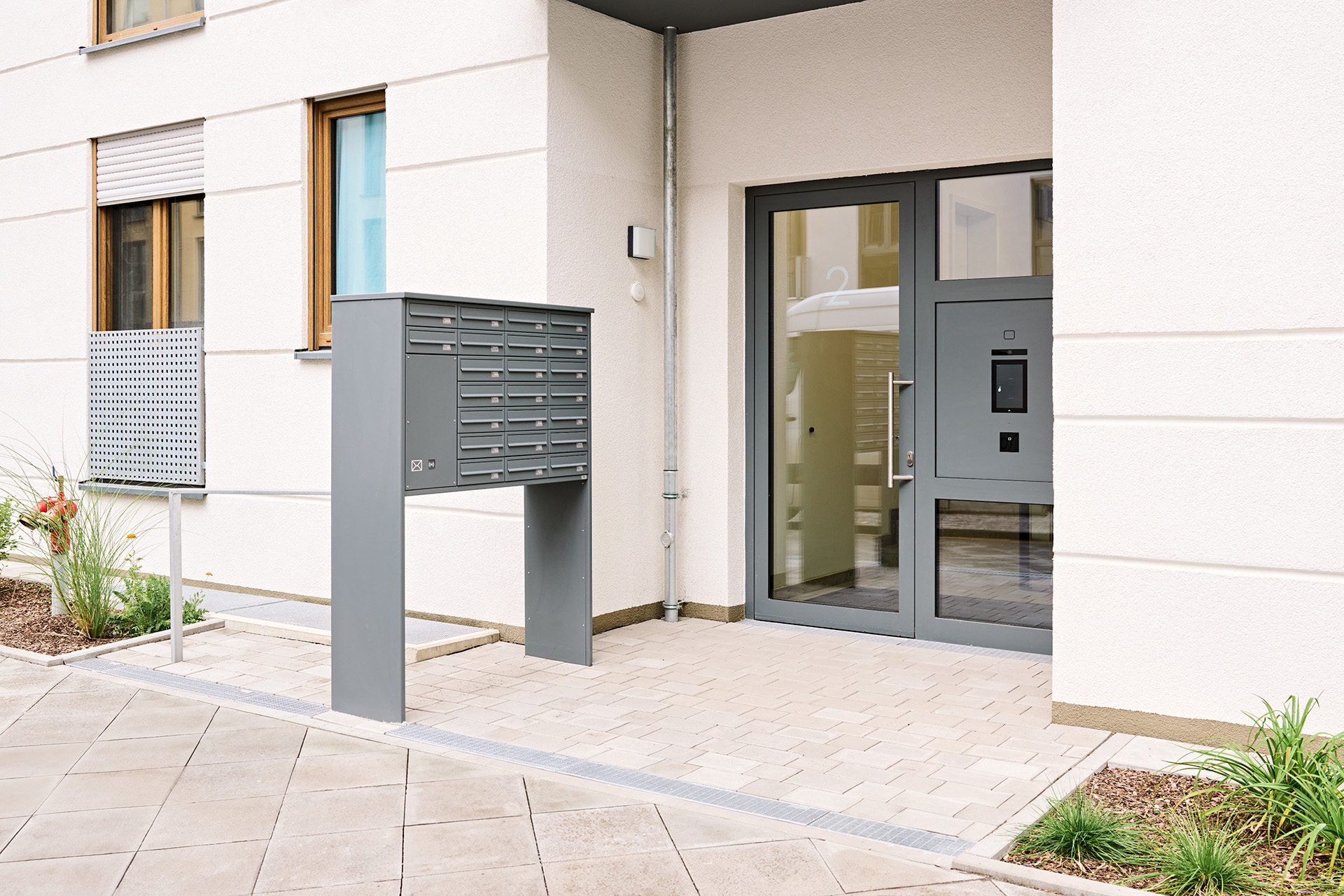 Mehrere graue Briefkästen vor dem Eingang eines hellen Wohnhauses mit Glastür und Sprechanlage.