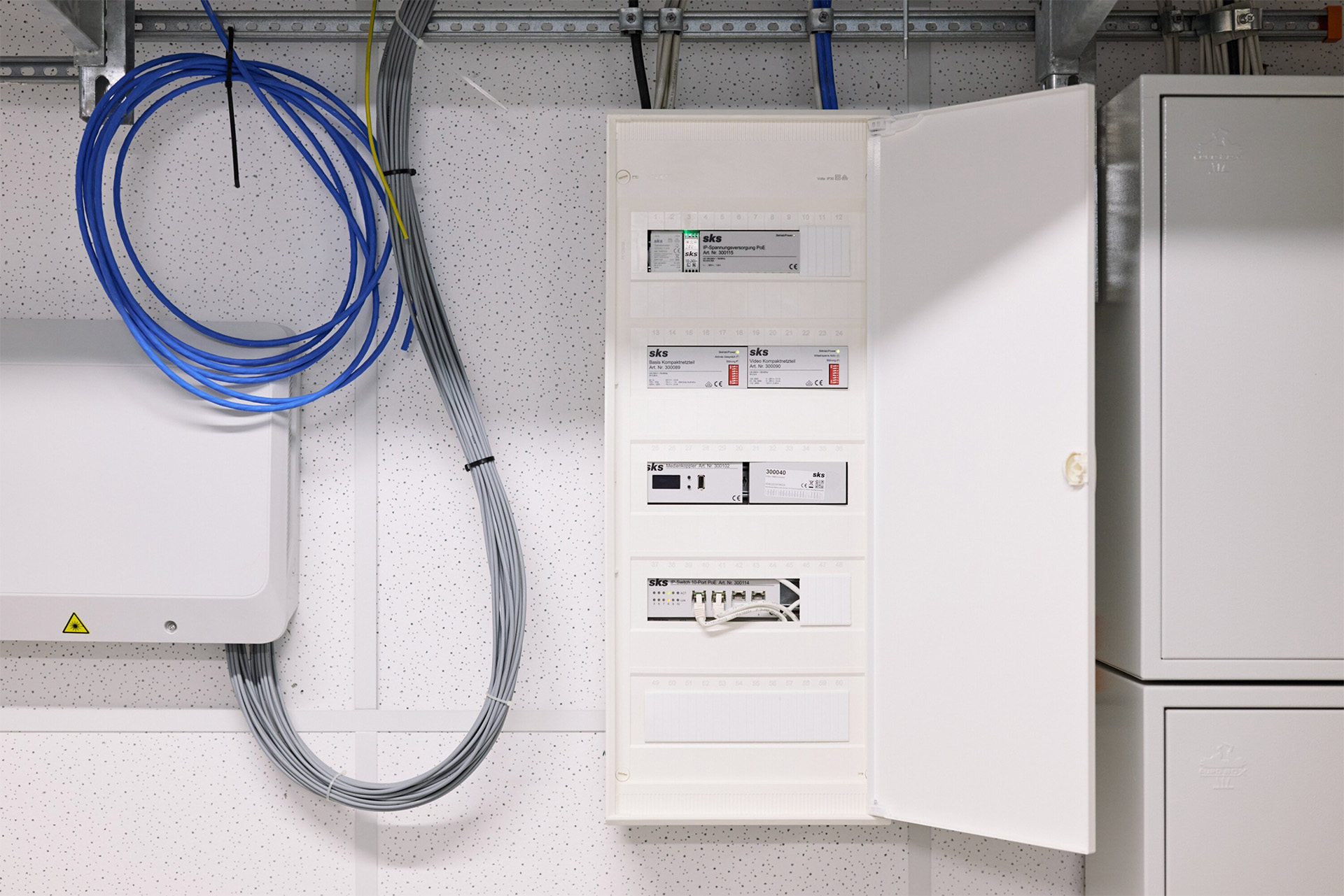 Geöffneter weißer Sicherungskasten mit mehreren Schaltern und Zählern, daneben Kabel in blauer und grauer Ummantelung an der Wand befestigt.