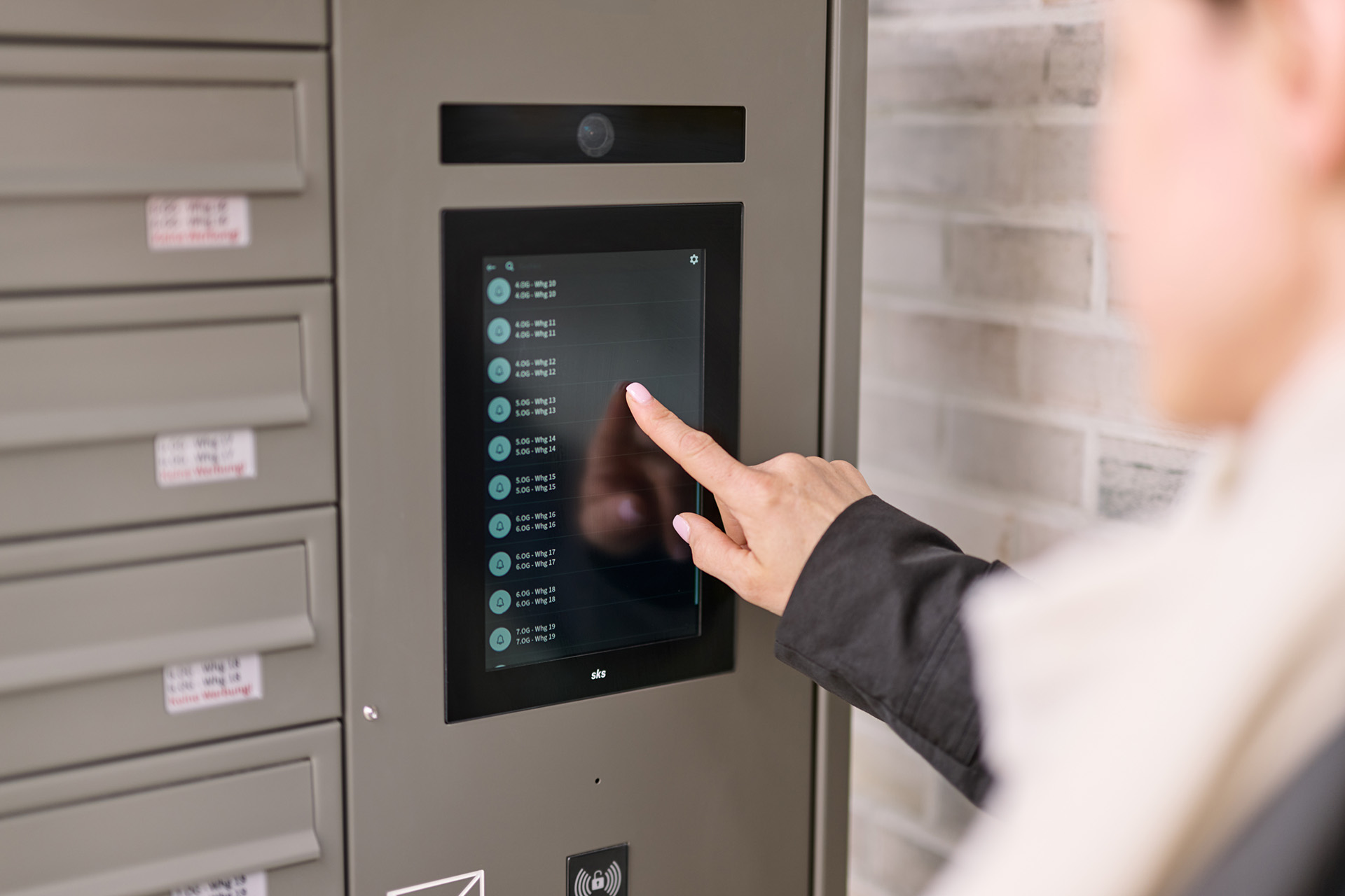 Hand tippt auf Touchscreen eines schwarzen Klingelsystems mit beleuchteter Benutzeroberfläche, links daneben Briefkästen