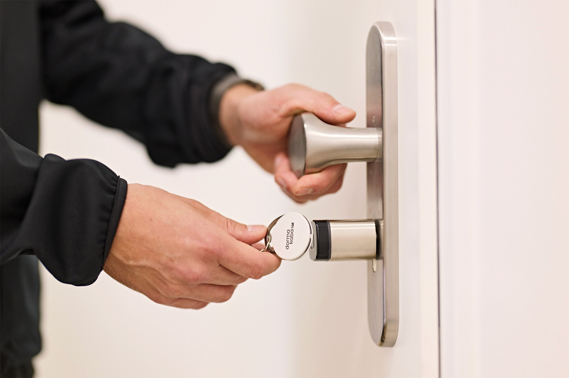 Hände hält einen runden Schlüsselanhänger mit dem Schriftzug 'dormakaba' gegen ein elektronisches Türschloss mit Griff.