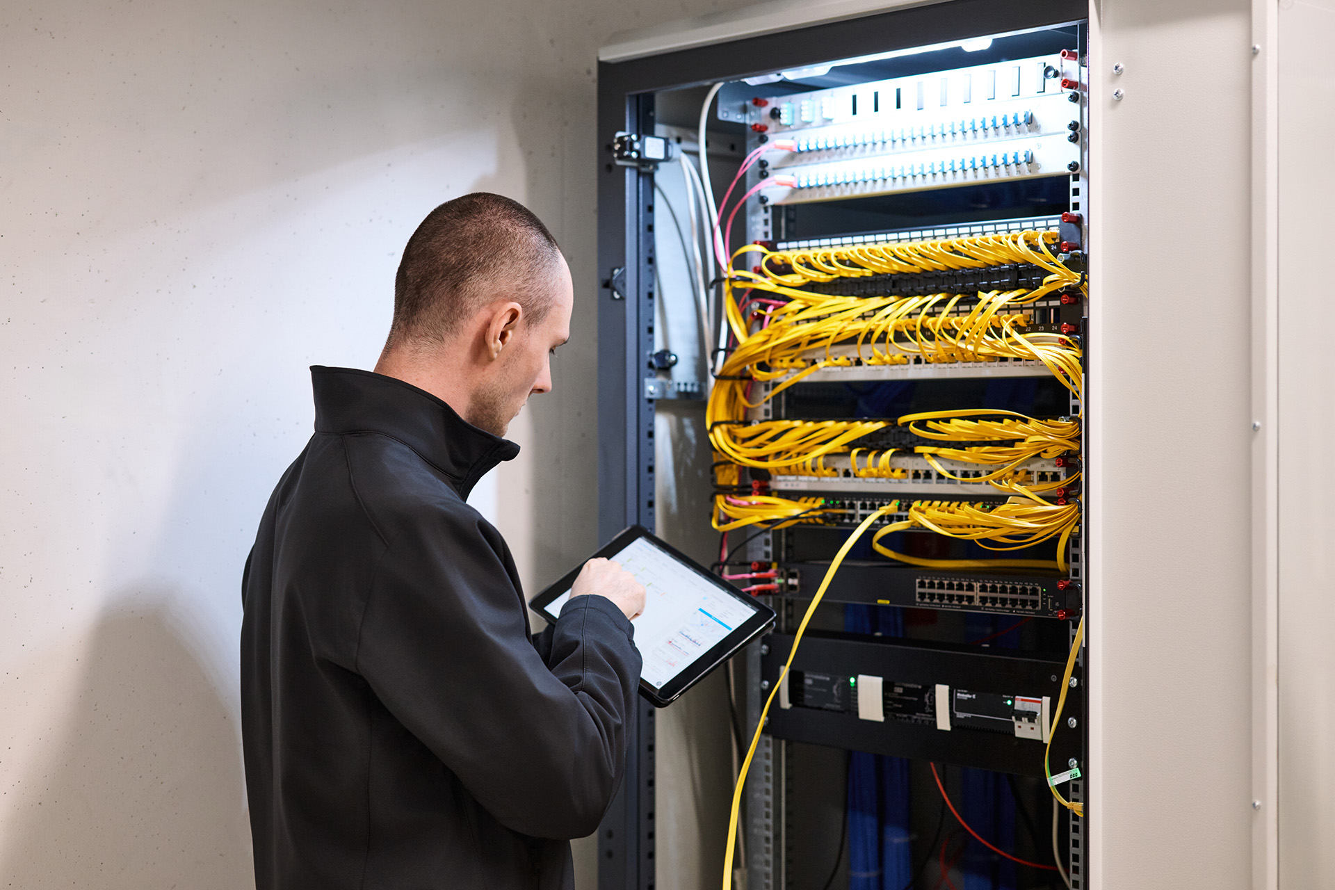 Person in schwarzer Jacke steht vor einem offenen Serverschrank mit zahlreichen gelben Netzwerkkabeln und hält ein Tablet in der Hand.