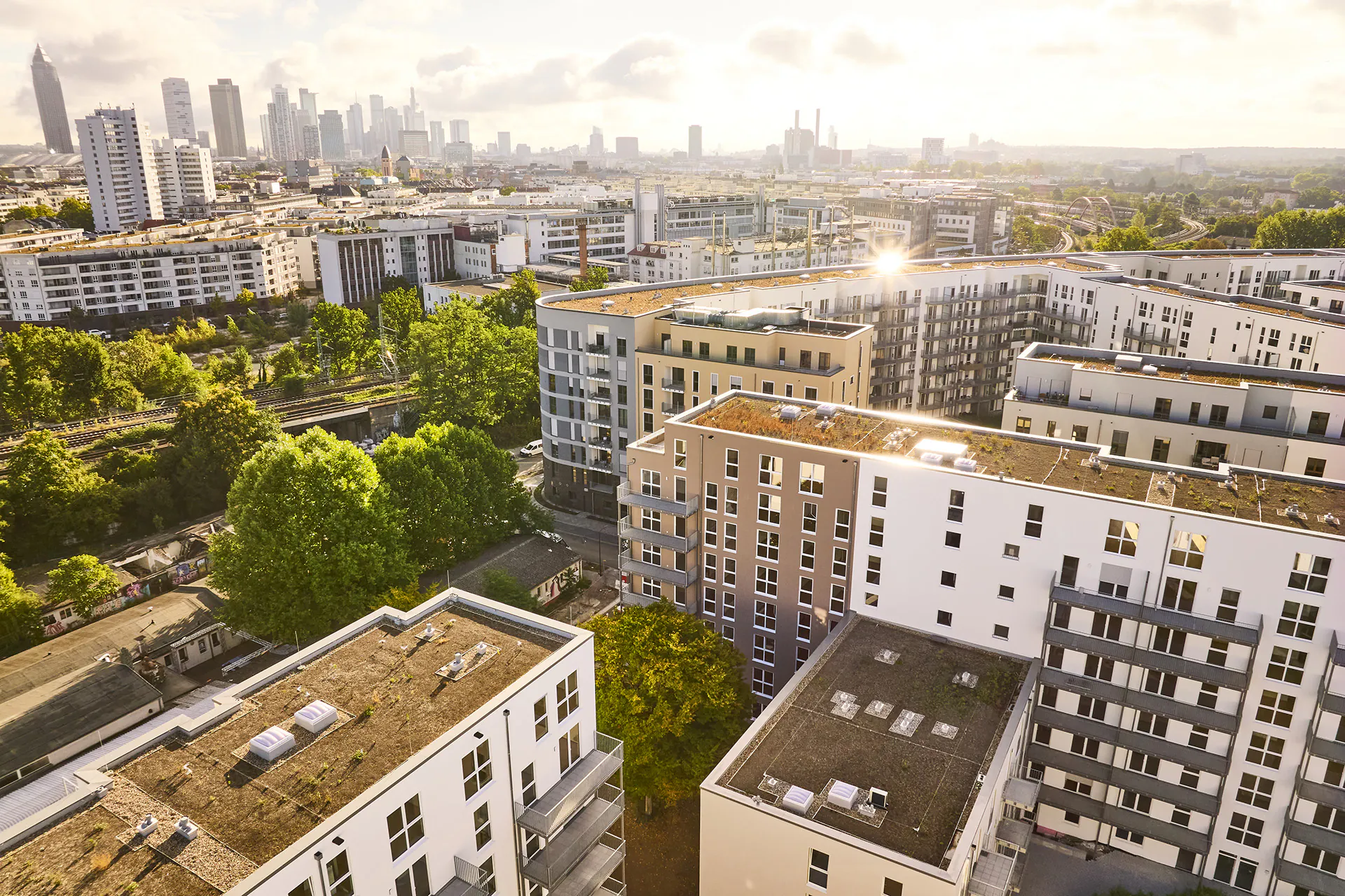 Luftaufnahme eines Wohngebiets mit mehrstöckigen Gebäuden, begrünten Innenhöfen und einer Stadt im Hintergrund bei Sonnenlicht.