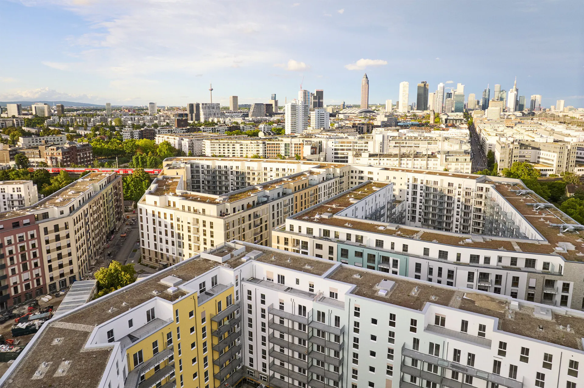 Luftaufnahme von Wohngebäuden mit Flachdächern und der Skyline einer Großstadt im Hintergrund bei klarem Himmel.